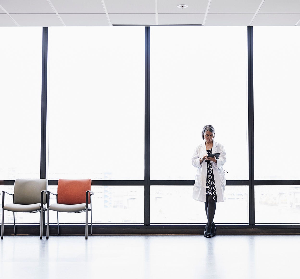 A woman in a lab coat leans again a large office while on a tablet in an empty waiting room.